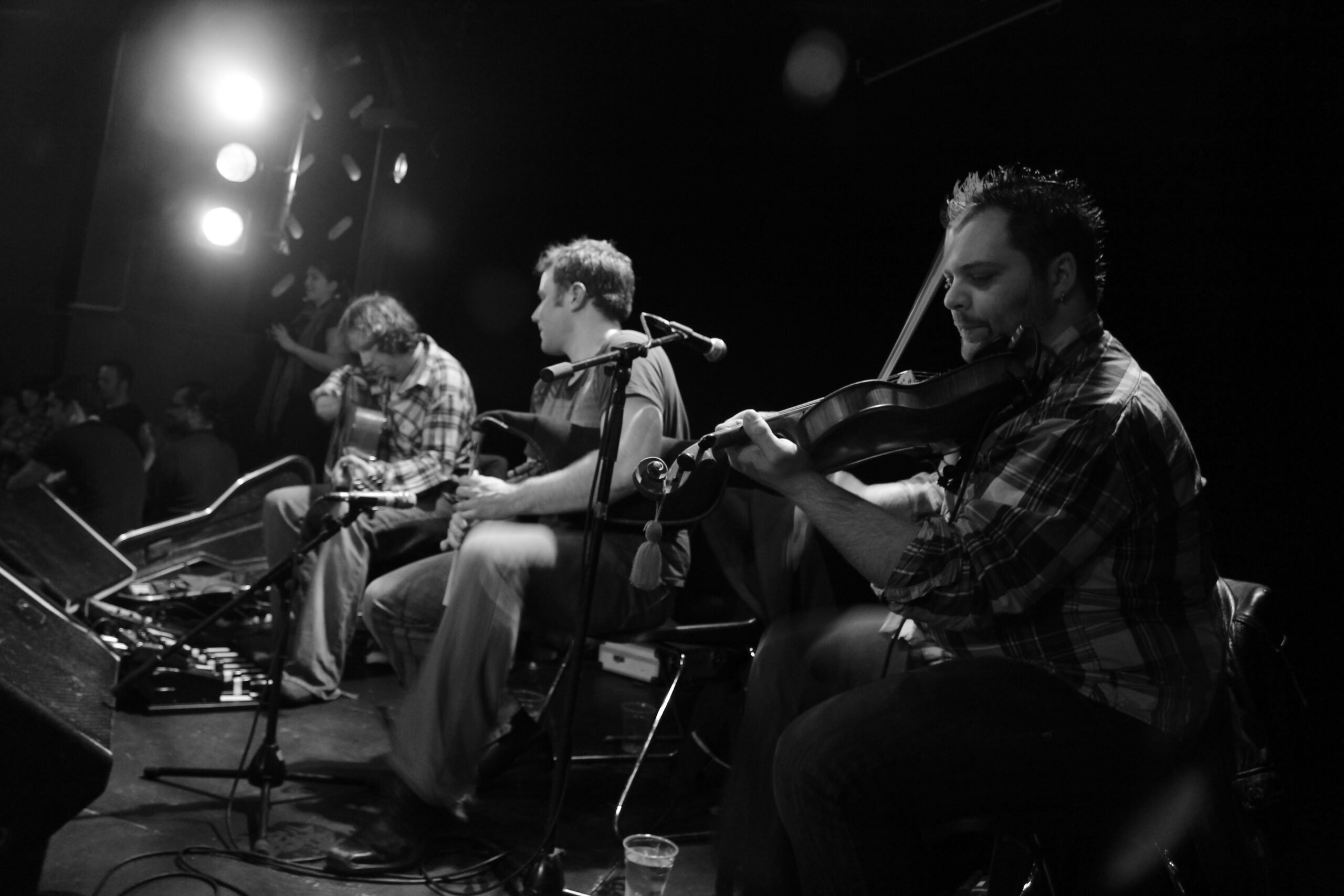 The Or Ceilidh band on stage photographed in black and white. Alex Urqhuart-Taylor features in the center of the image playing bagpipes. A guitarist and a fiddler also feature in the image