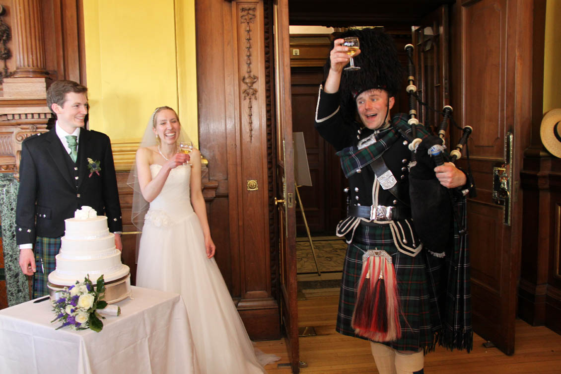 A photo of a piper in full kilt and highland dress, holding bagpipes and toasting a married couple on their wedding day.